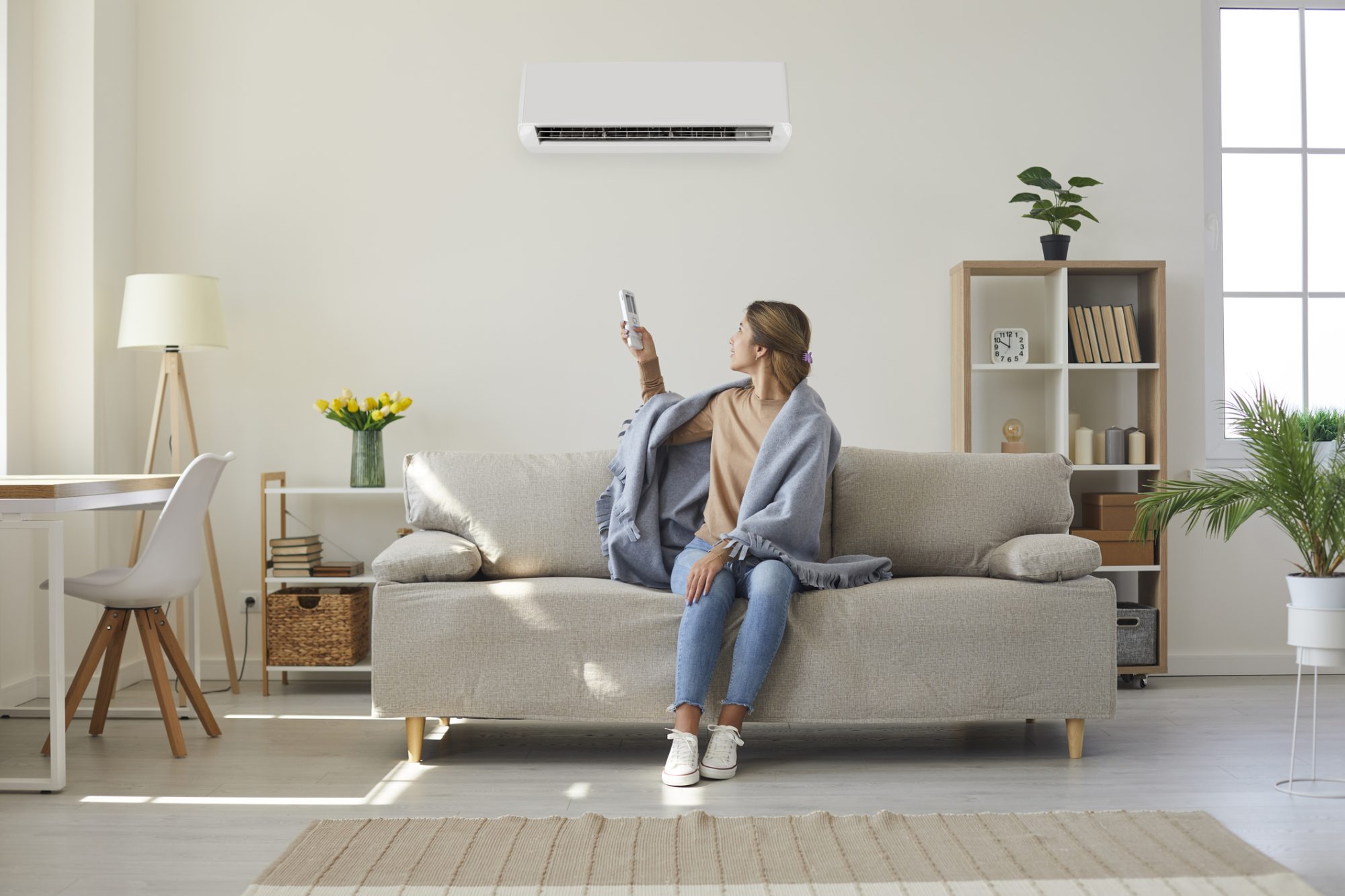 Woman enjoying cool fresh air in her living room with air conditioner on the wall. Woman who& x27;s sitting on sofa under warm plaid in living room switches off her air conditioner on wall. Young girl adjusting modern AC system, regulating temperature and enjoying cool fresh air at home